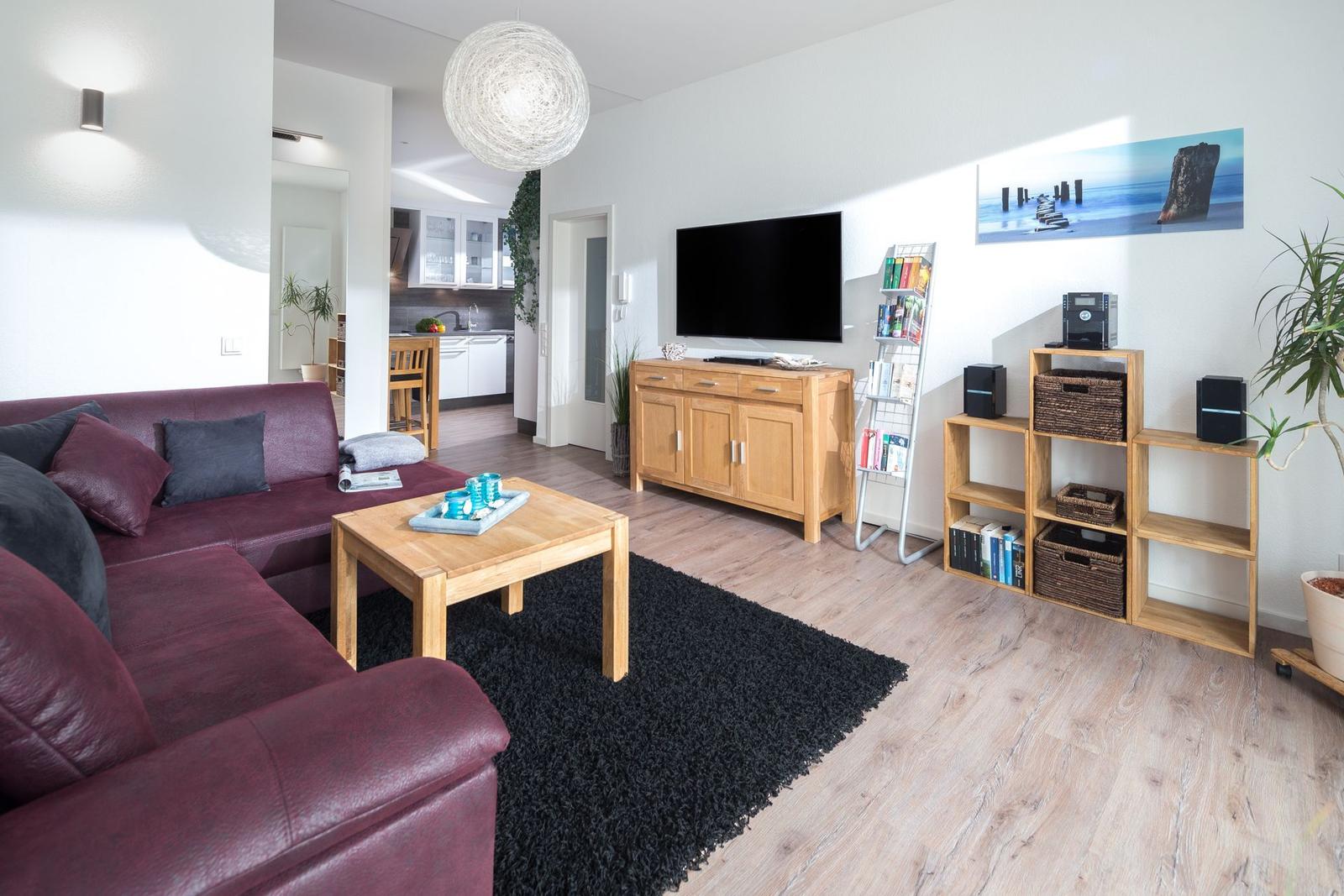 Wohnzimmer mit Sofa, Couchtisch und Fernseher. Küche im Hintergrund sichtbar.