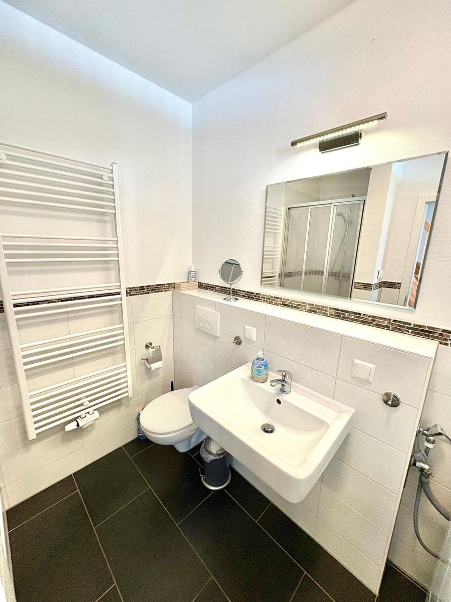 Badezimmer mit Toilette, Waschbecken und Dusche. Weiße Wände, dunkler Boden und Heizung.