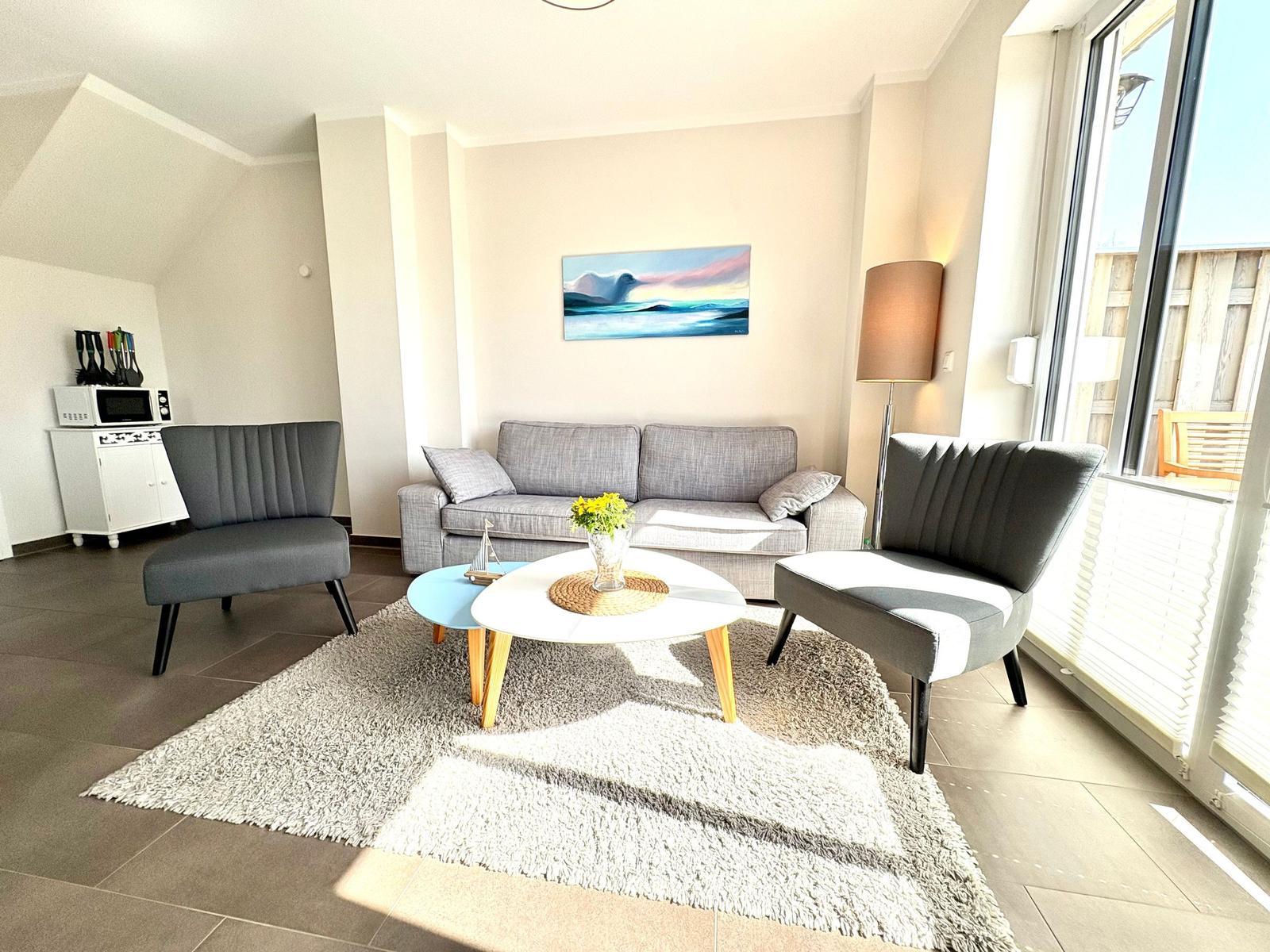 Wohnzimmer mit Sofa, zwei Sesseln und Tisch. Fenster zum Balkon. Wandbild und Stehlampe.