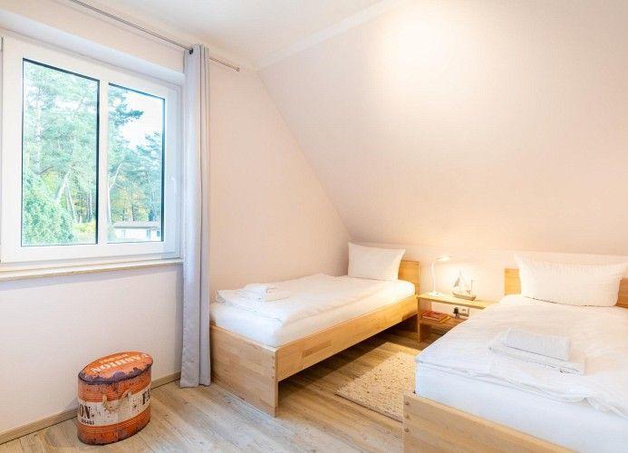 Zwei Betten mit weißer Bettwäsche, Fenster mit Blick auf Wald, Holzboden und kleiner Sitzhocker.