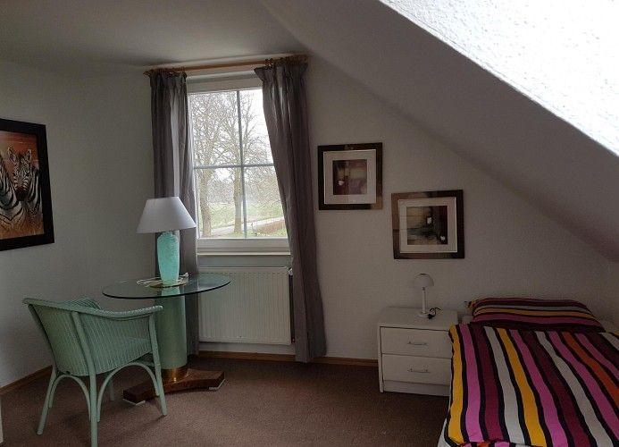 Schlafzimmer mit Dachschräge, Bett, Nachttisch, Stuhl und Tisch unter Fenster.