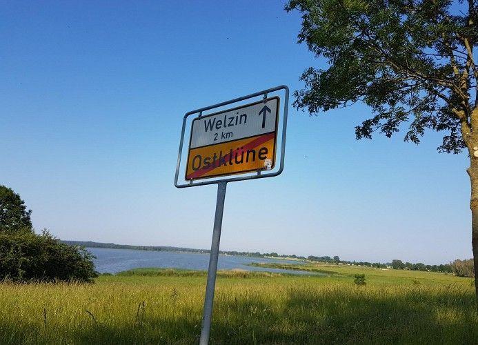 Schild weist auf Welzin und Ostklüne hin, mit Blick auf Wasser und Grasland.
