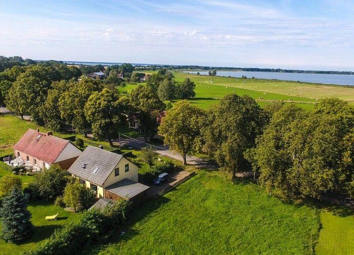 Haus mit rotem Dach und gelbem Anbau, umgeben von Grünflächen und Bäumen, nahe einem See.