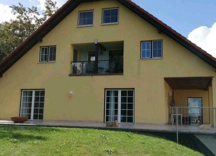 Haus mit Balkon und Terrasse, umgeben von Grünfläche und Bäumen.