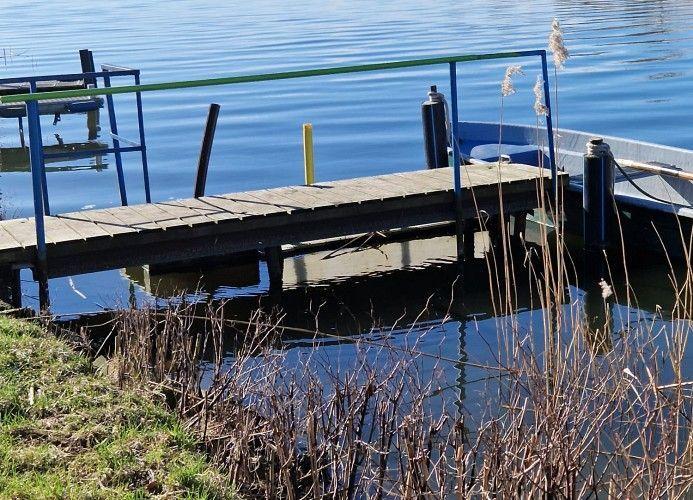 Holzsteg mit Geländer führt zum Wasser. Im Hintergrund liegt ein Boot.