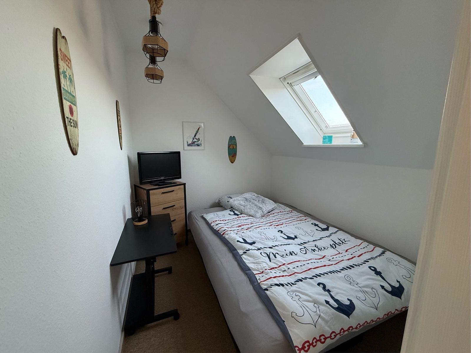 Schlafzimmer mit Dachschrägen, Bett mit Ankern-Motiv, TV und Schreibtisch.