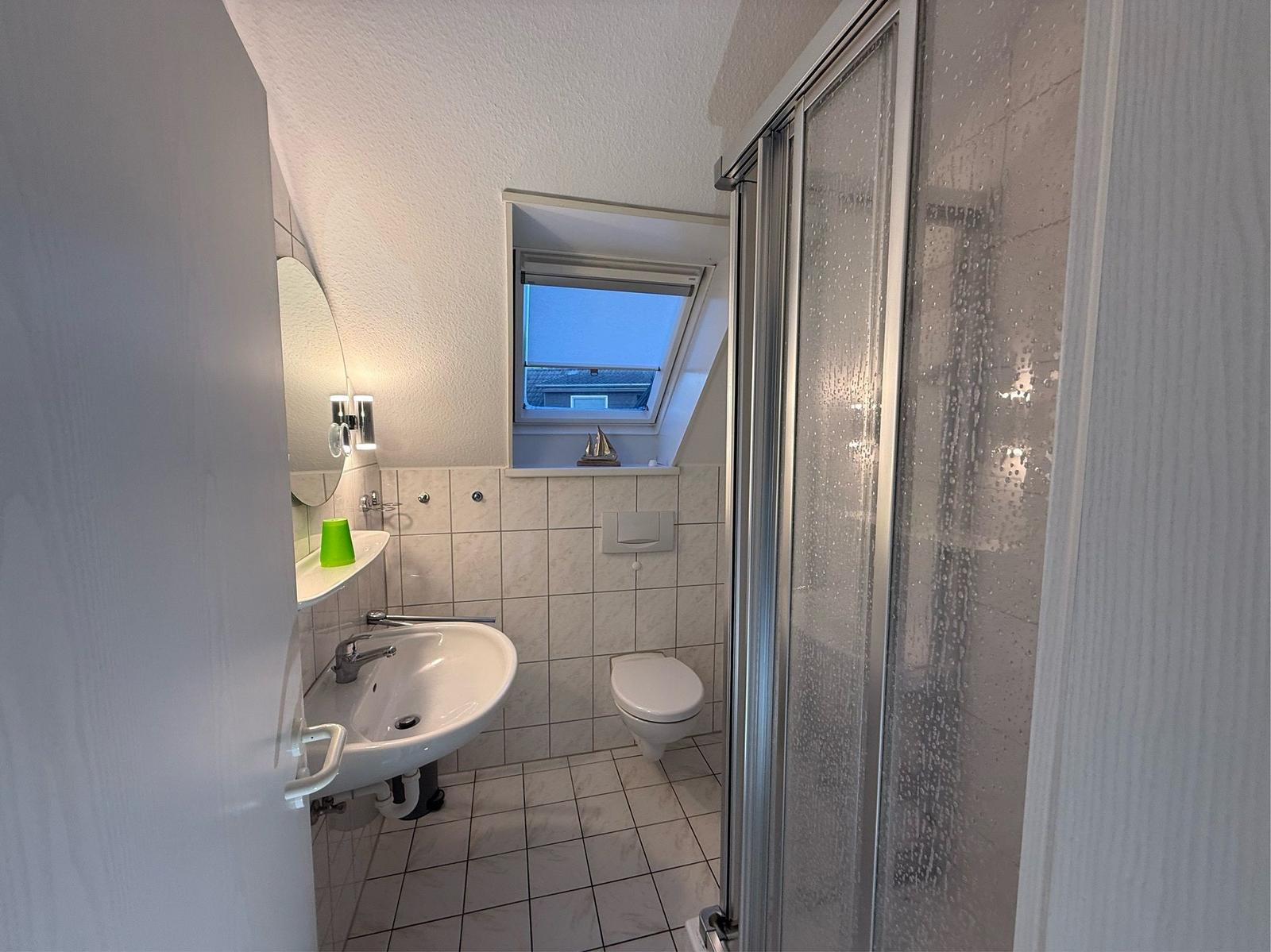 Badezimmer mit Dusche, Waschbecken und Toilette. Dachfenster mit Segelbootdekoration.