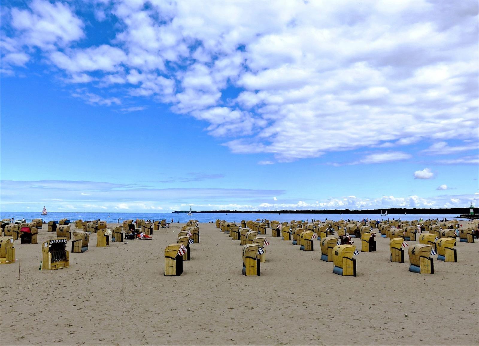 Strand mit gelben Strandkörben unter blauem Himmel mit Wolken und Booten im Wasser.