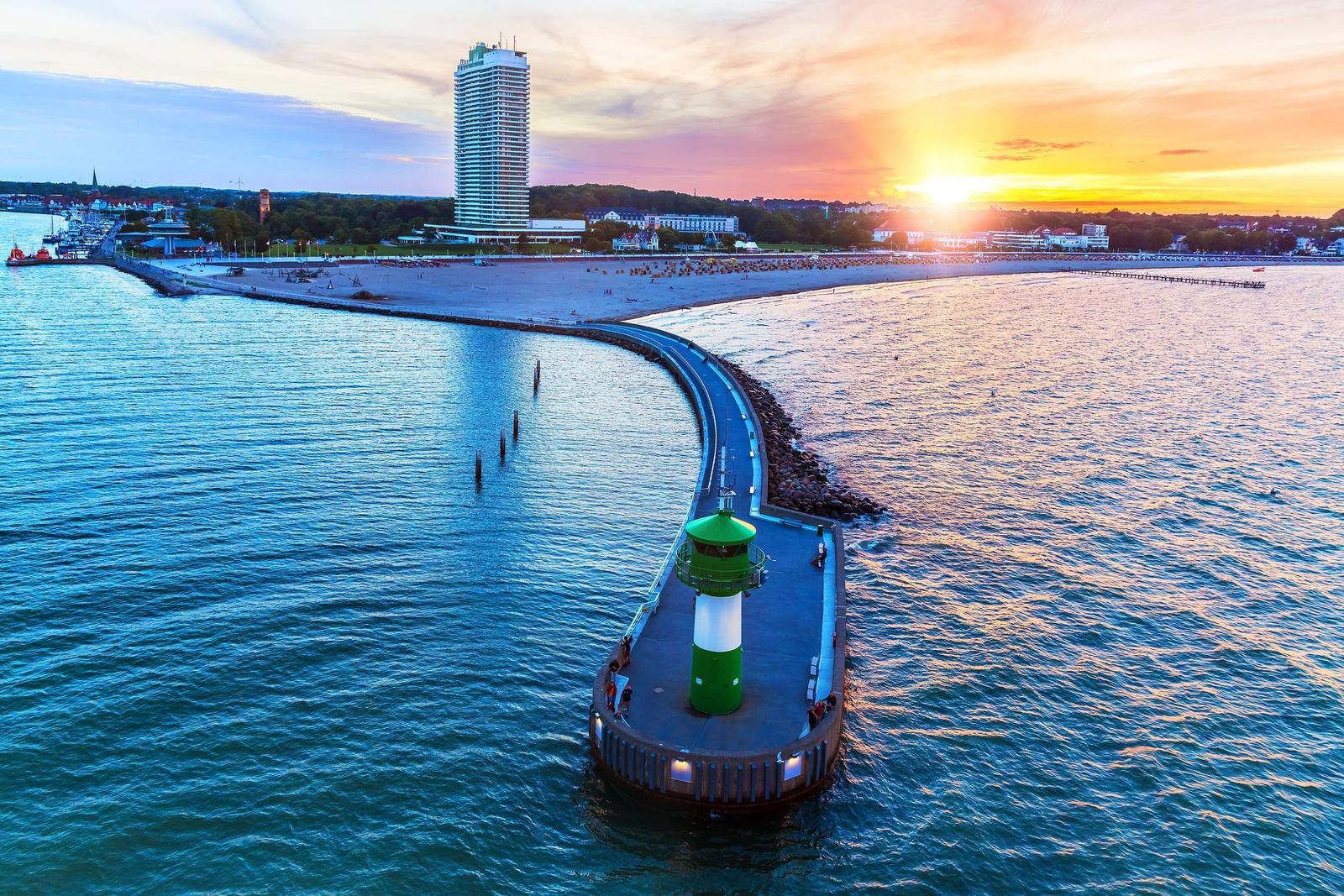Hafenbecken mit Leuchtturm und Strand bei Sonnenuntergang.