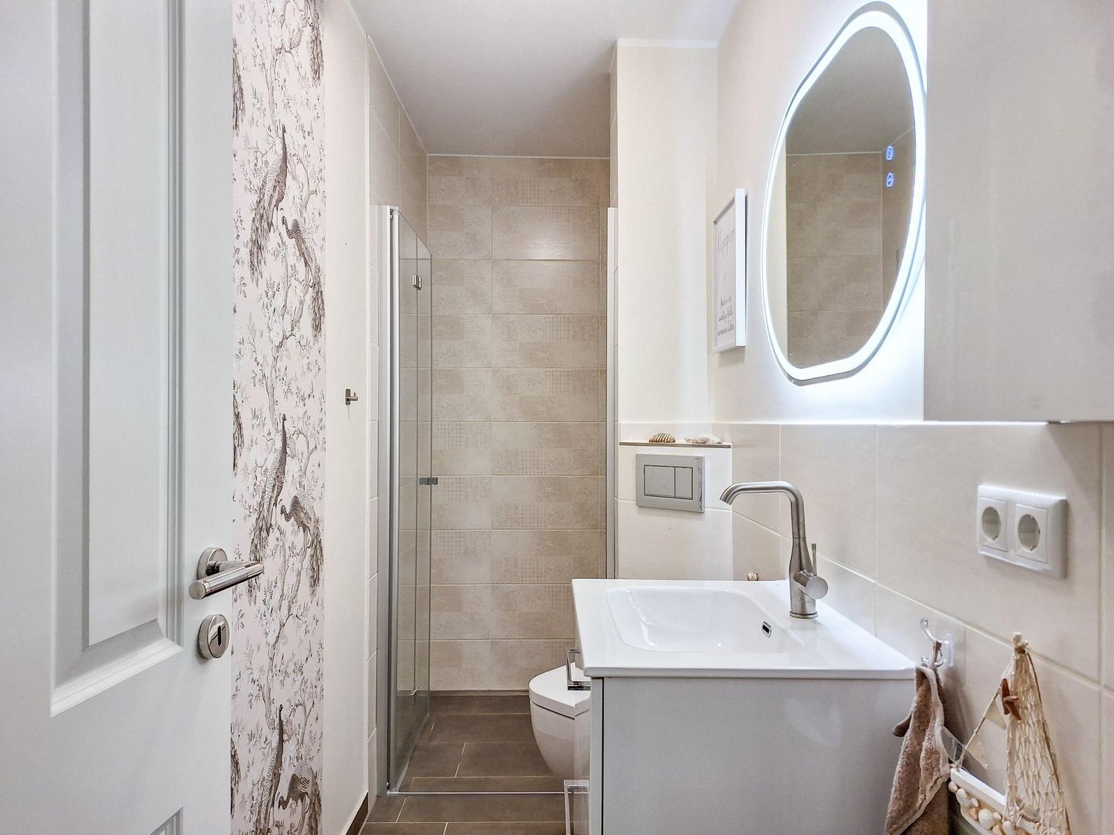 Badezimmer mit Dusche, Waschbecken und Toilette. Wand mit Tapete und beleuchtetem Spiegel.