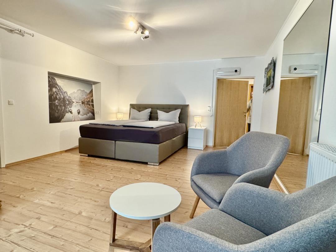 Schlafzimmer mit Bett, Sessel und Wandbild. Holzboden und Deckenleuchten.