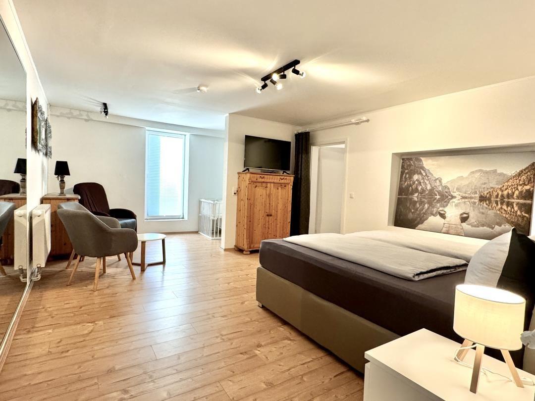 Schlafzimmer mit Bett, Fernseher und Wandbild. Holzboden und Sessel im Raum.