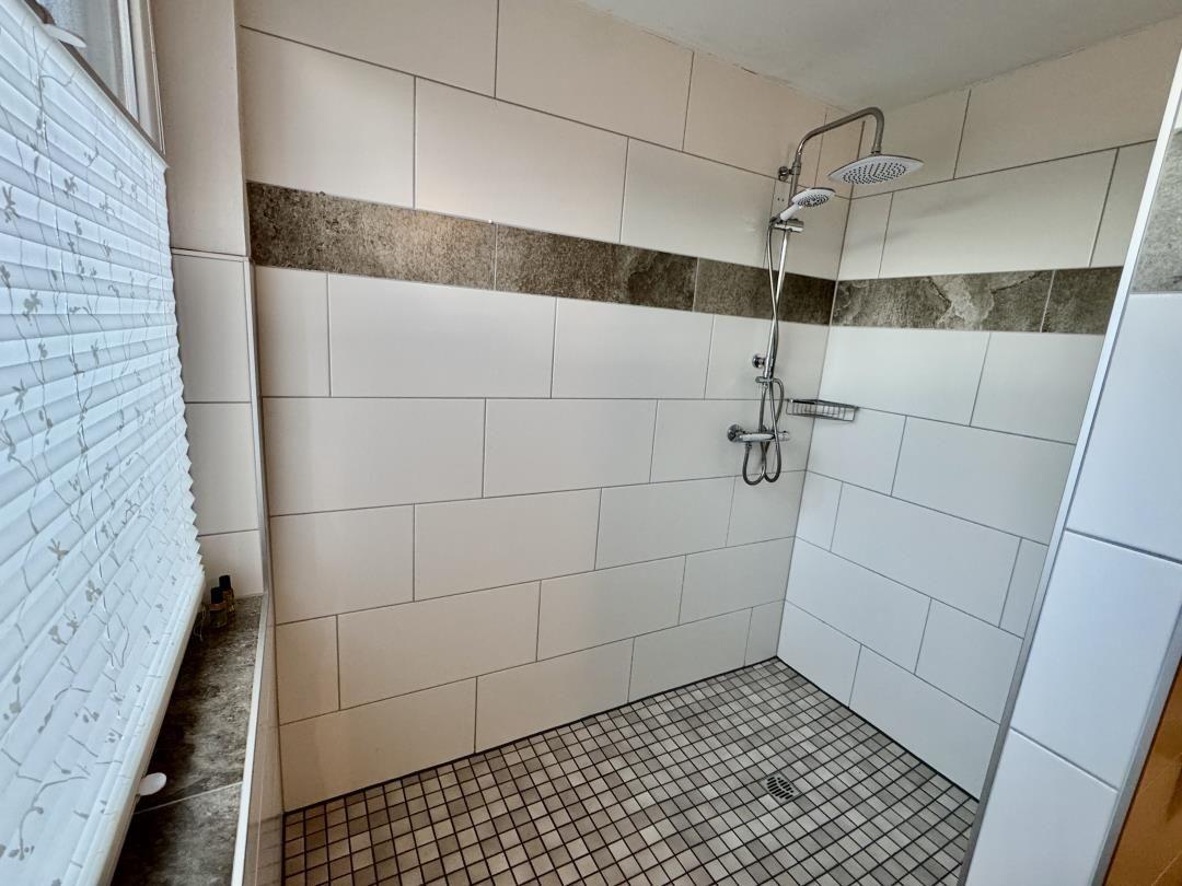 Dusche mit weißen Fliesen und dunkler Zierleiste. Bodenfliesen und Duschbrause sichtbar.