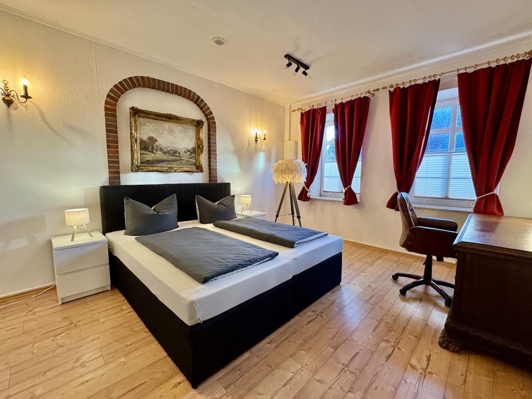 Schlafzimmer mit Doppelbett, Schreibtisch, roten Vorhängen und Holzboden.