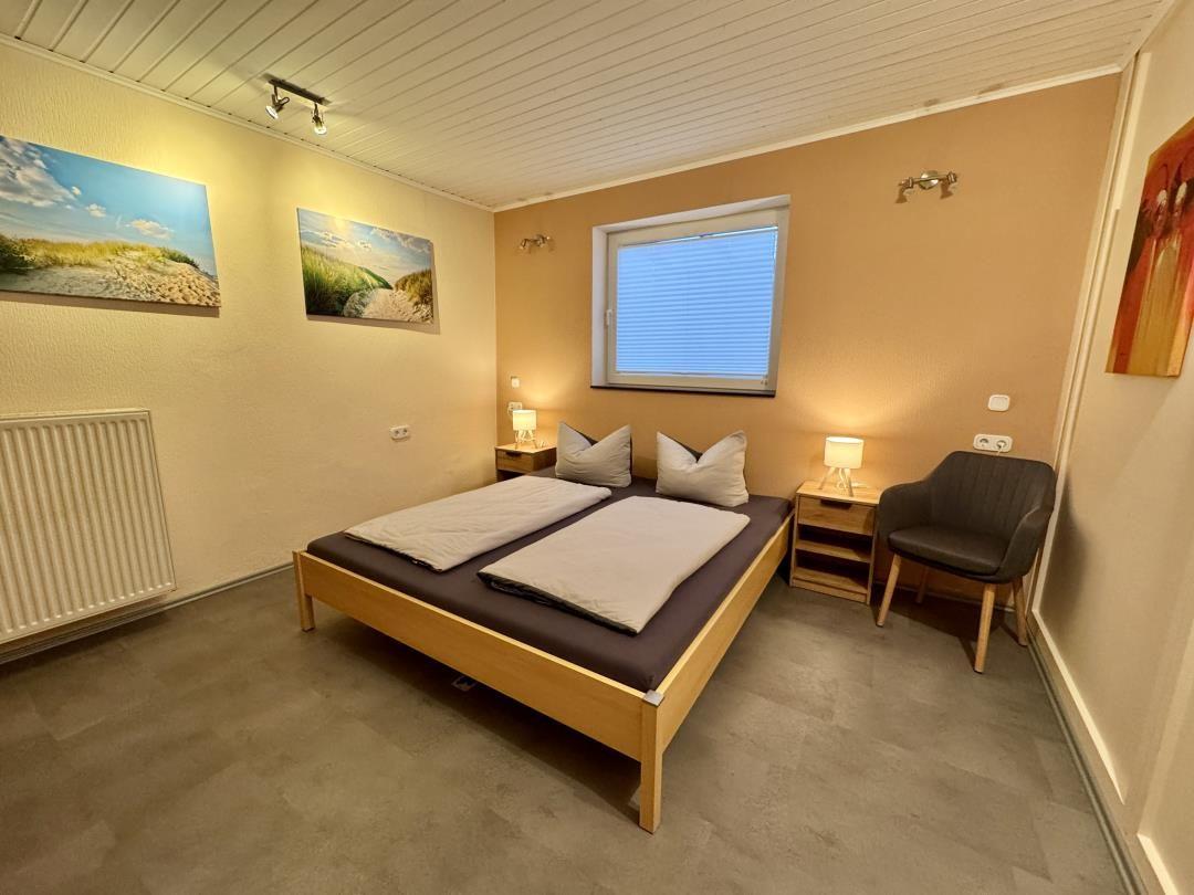 Schlafzimmer mit Doppelbett, zwei Nachttischen, Sessel und Fenster mit Jalousie.