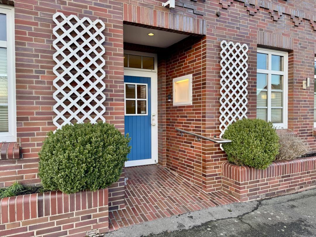 Eingang mit blauer Tür, rotem Backsteinhaus und weißen Gitterelementen.