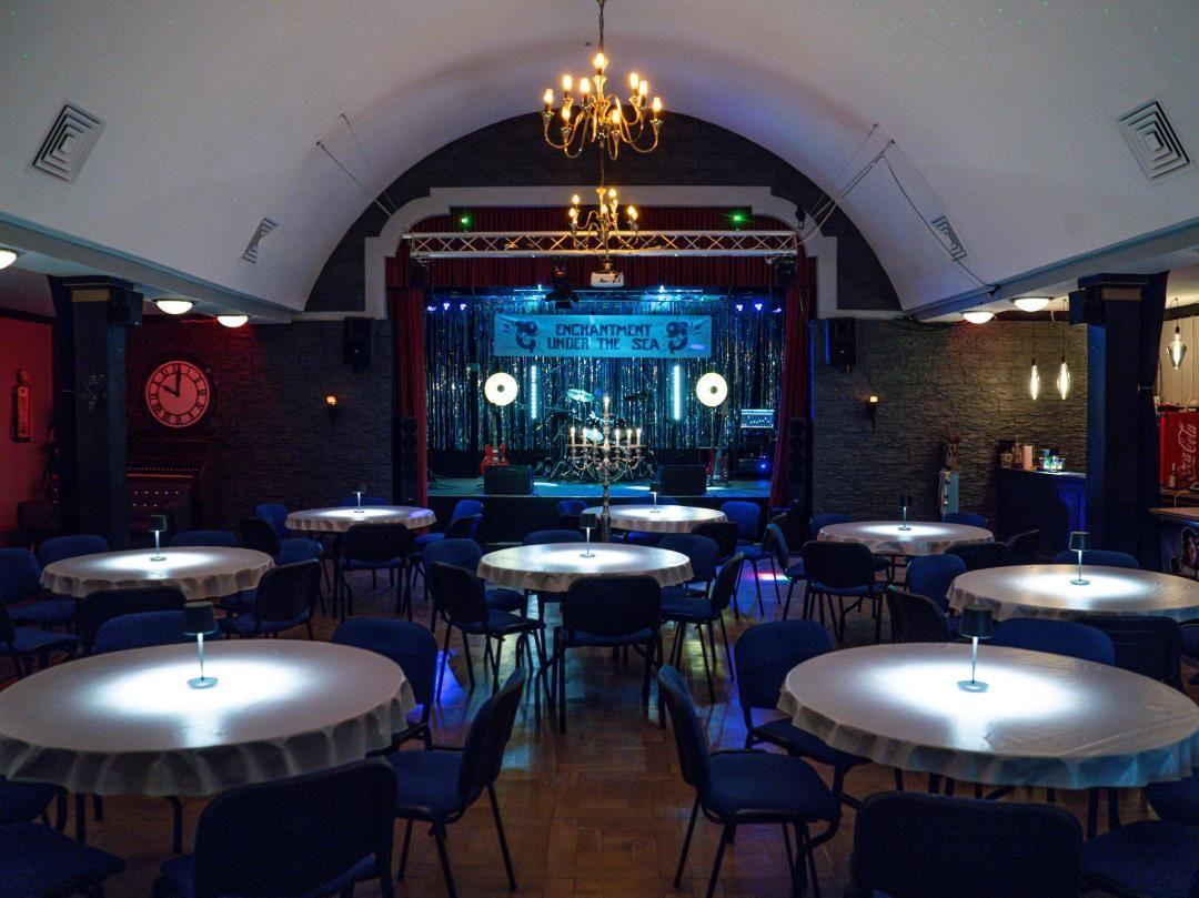 Großer Saal mit runden Tischen, Bühne und Kronleuchter. Bühnenaufbau für Veranstaltungen.