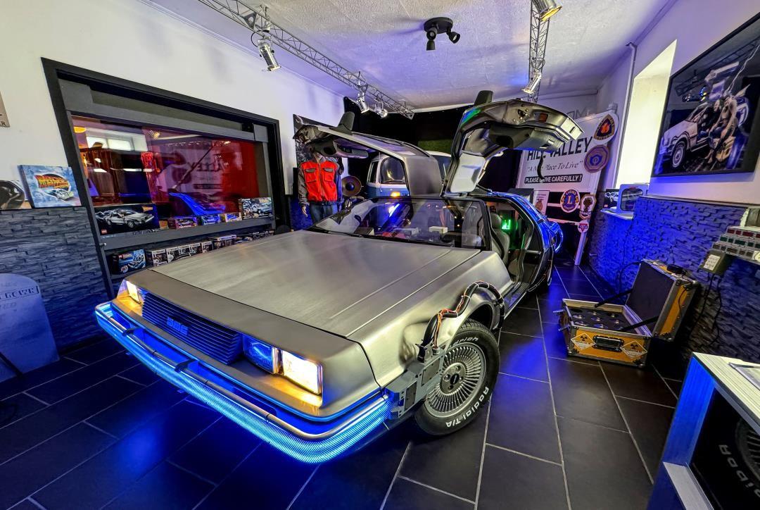Ein silberner DeLorean mit geöffneten Türen steht in einem Raum mit blauer Beleuchtung.