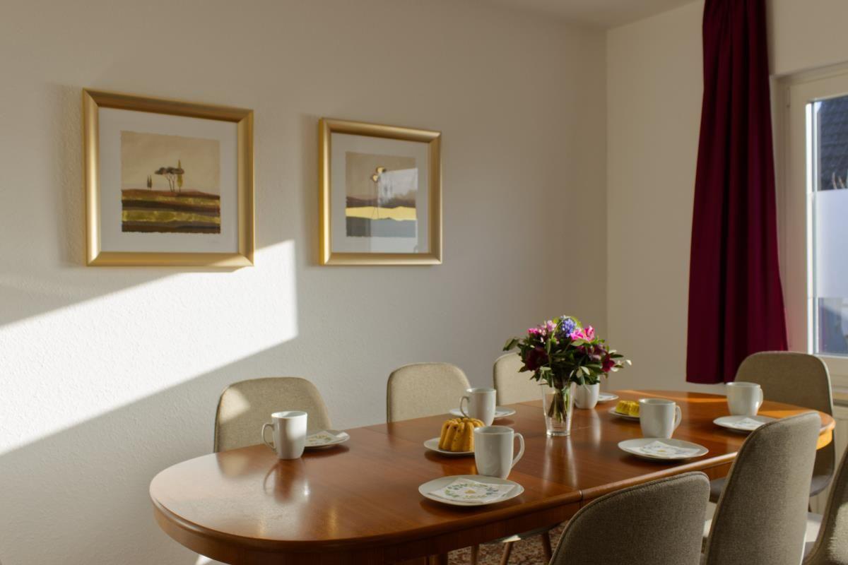 Esstisch mit Stühlen, Tassen, Teller und Blumenarrangement an der Wand.