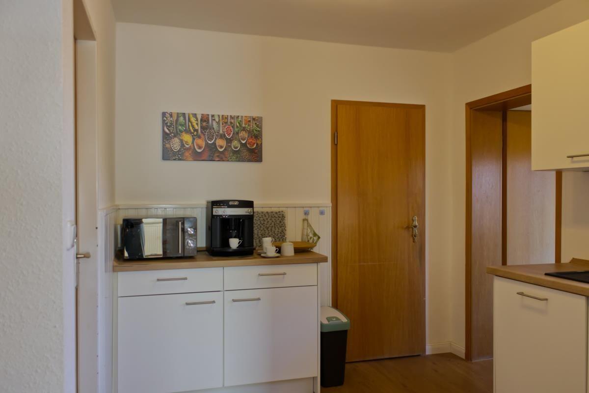 Küche mit Arbeitsplatte, Kaffeemaschine und Mikrowelle. Holztür und Wandbild sichtbar.
