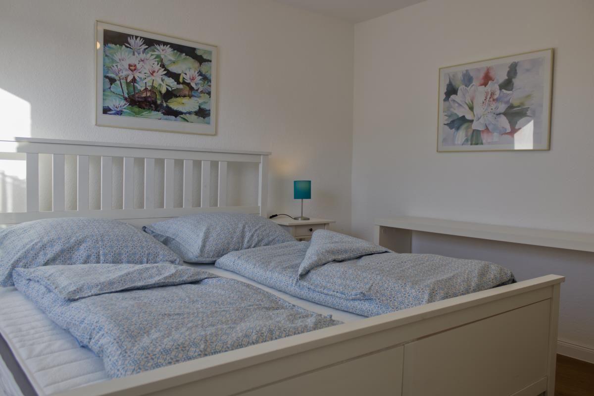 Schlafzimmer mit weißem Bett, zwei Bildern an der Wand und einer Nachttischlampe.