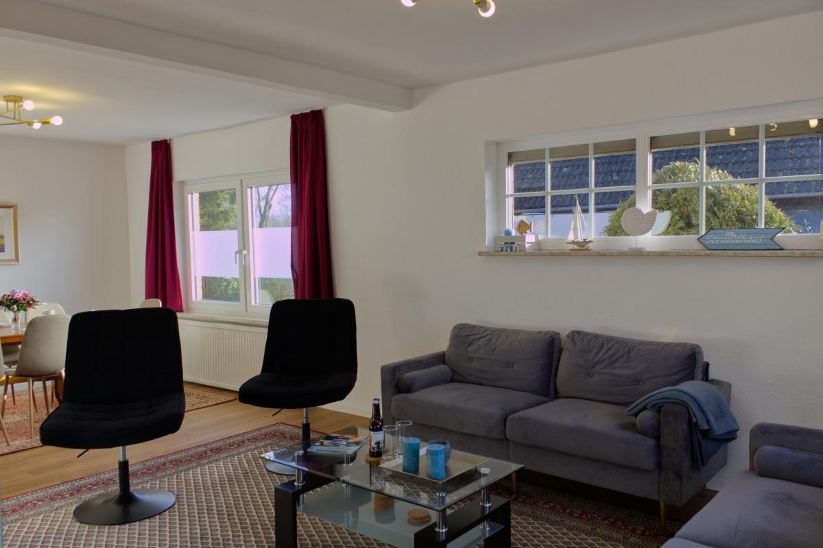 Wohnzimmer mit Sofa, zwei schwarzen Stühlen und Glas-Tisch. Fenster mit roten Vorhängen.