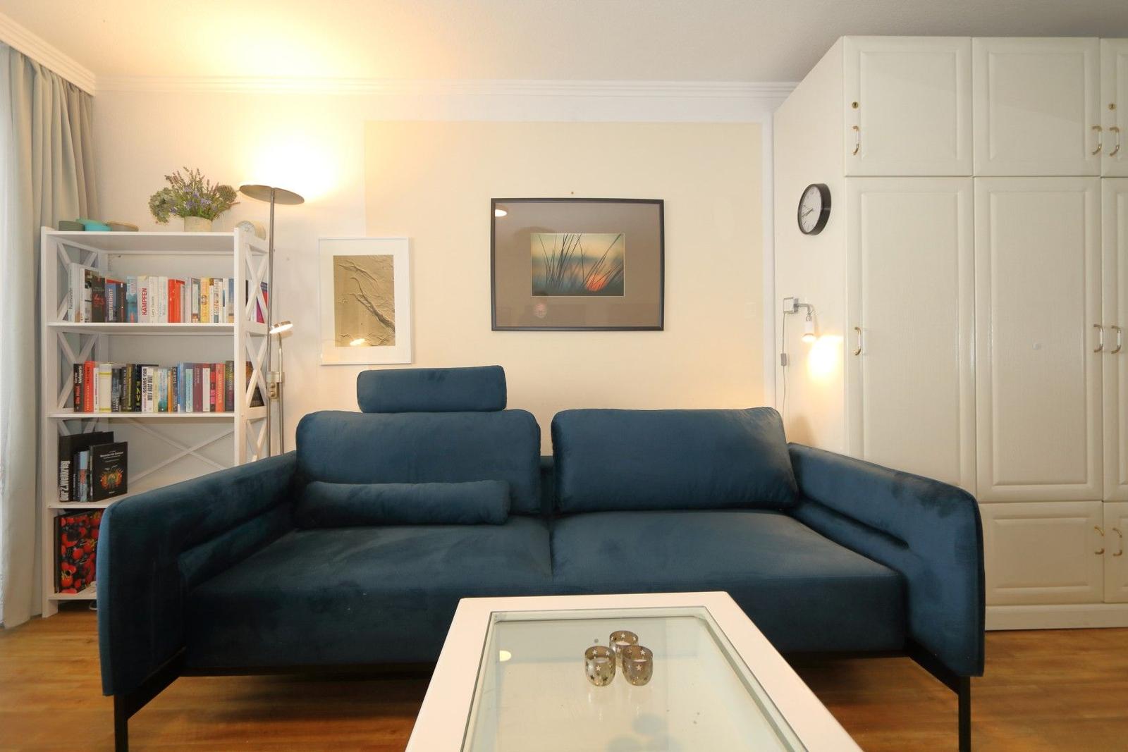 Ein Wohnzimmer mit blauem Sofa, Glas-Tisch und weißen Schränken an der Wand.