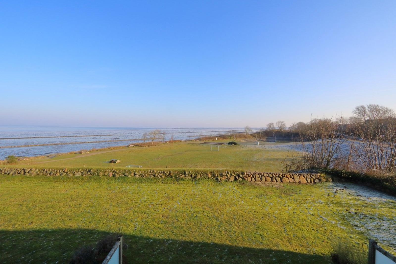 Green field with stone wall, view of sea and trees under blue sky.