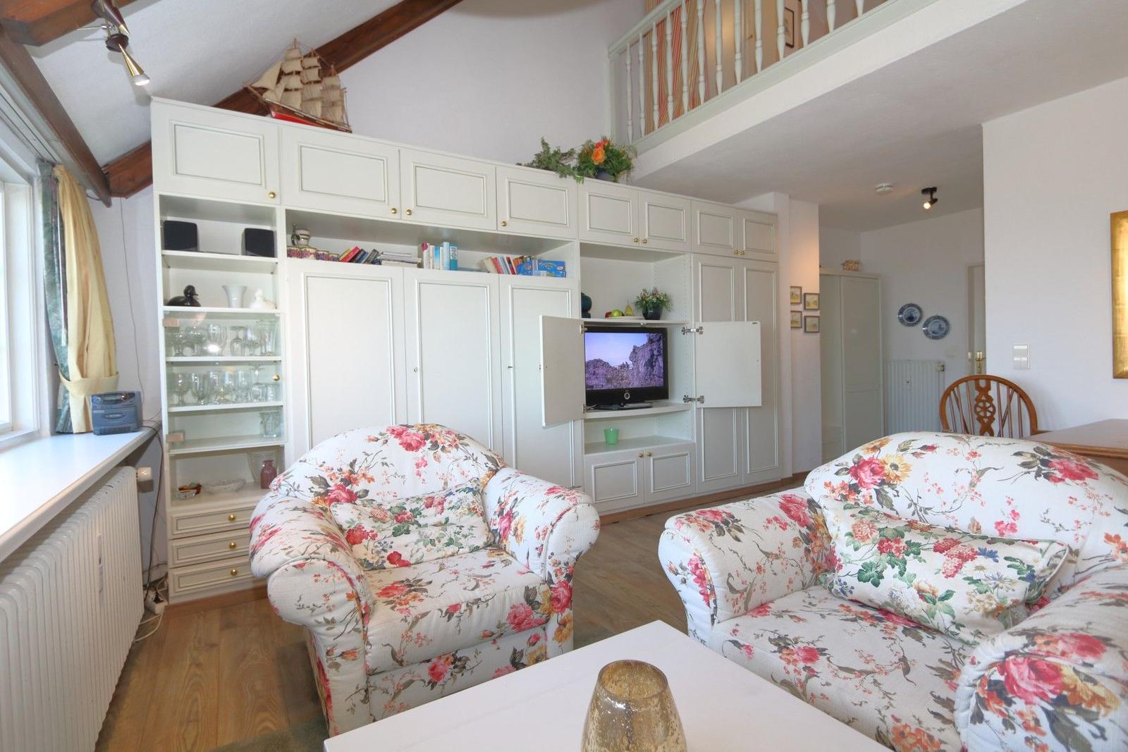 Living room with floral seating, TV cabinet, and balcony. Wooden floor and vaulted ceiling.