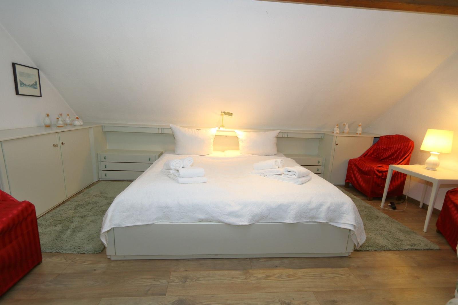 Bedroom with sloped ceilings, white bed, and red armchairs.