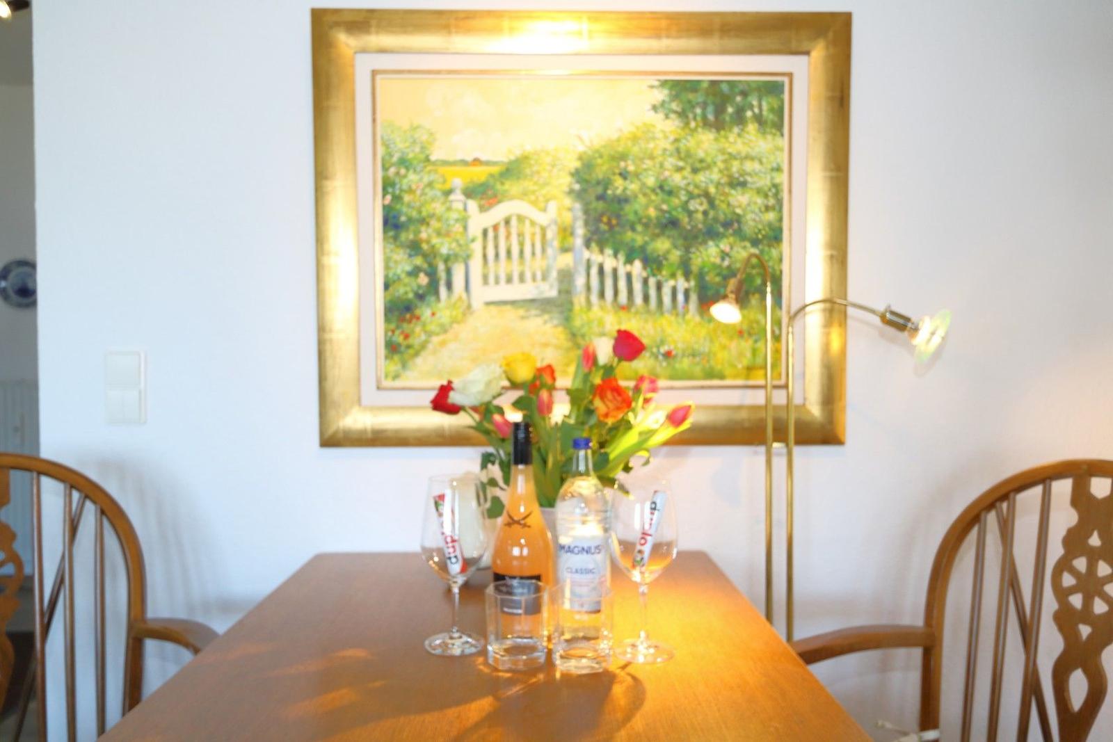 Dining table with flowers, bottles, and glasses in front of a garden-themed painting.