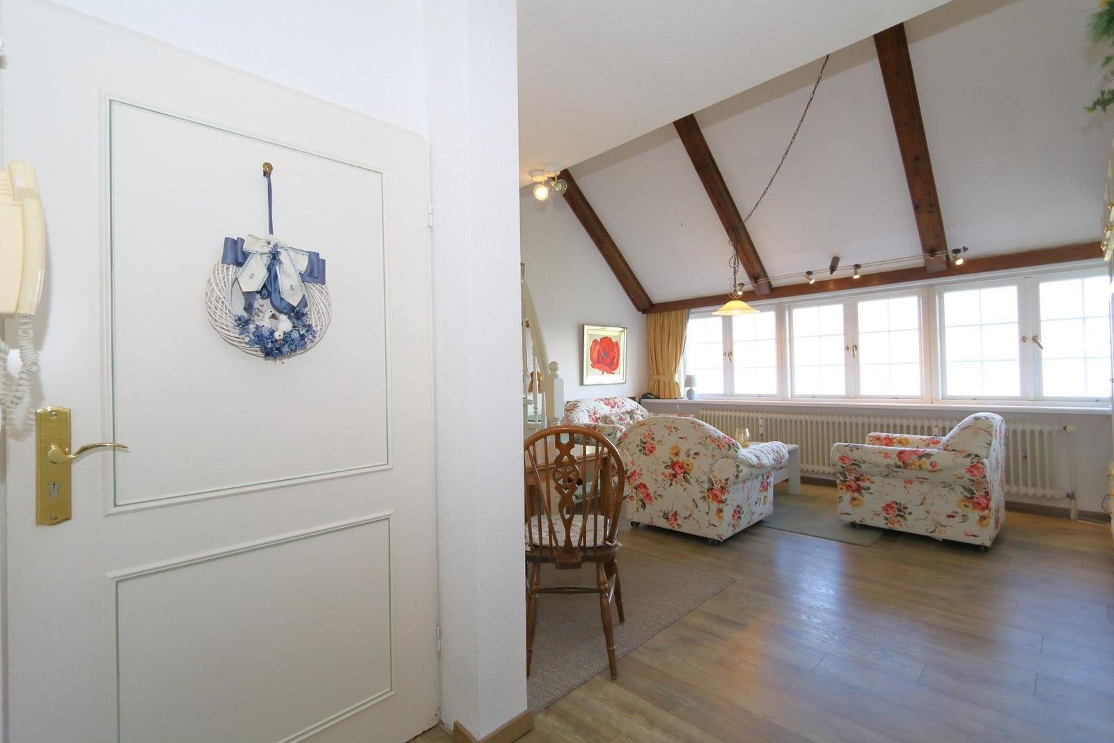 Entrance with door and view into living room with wooden beams and floral chairs.