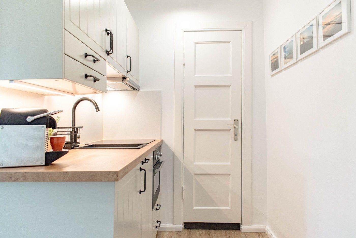 Küche mit Holzoberfläche, weißen Schränken und schwarzen Griffen. Weiße Tür und Wand mit Bildern.