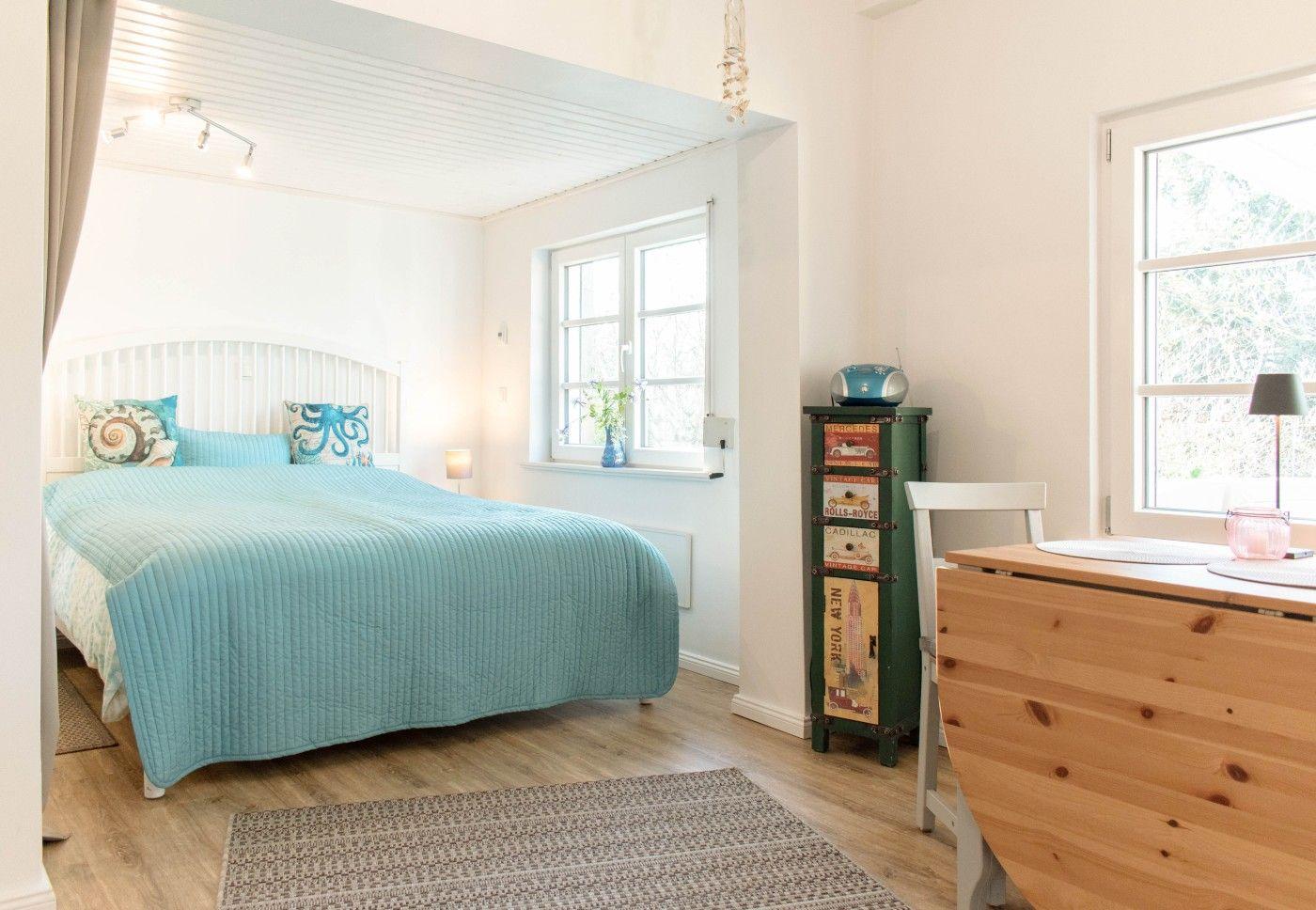 Schlafzimmer mit Bett, Schreibtisch und Fenstern. Holzboden und Wandfarbe weiß.