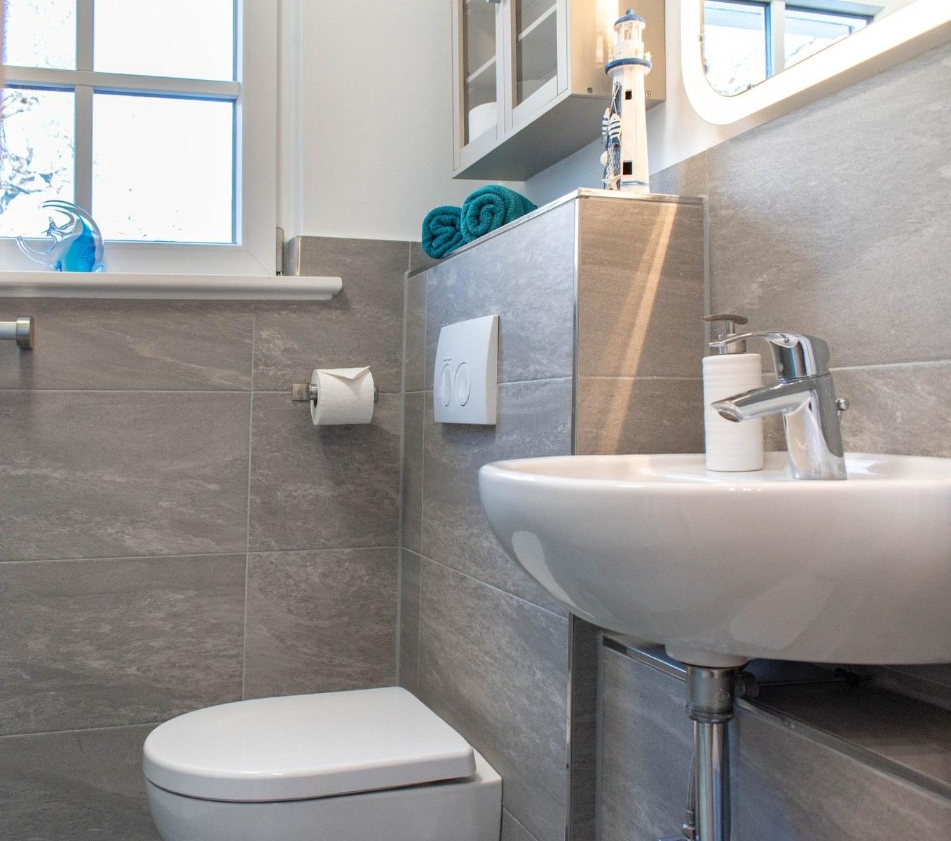 Badezimmer mit Toilette, Waschbecken und Fenster. Graue Fliesen und weißer Spiegel.