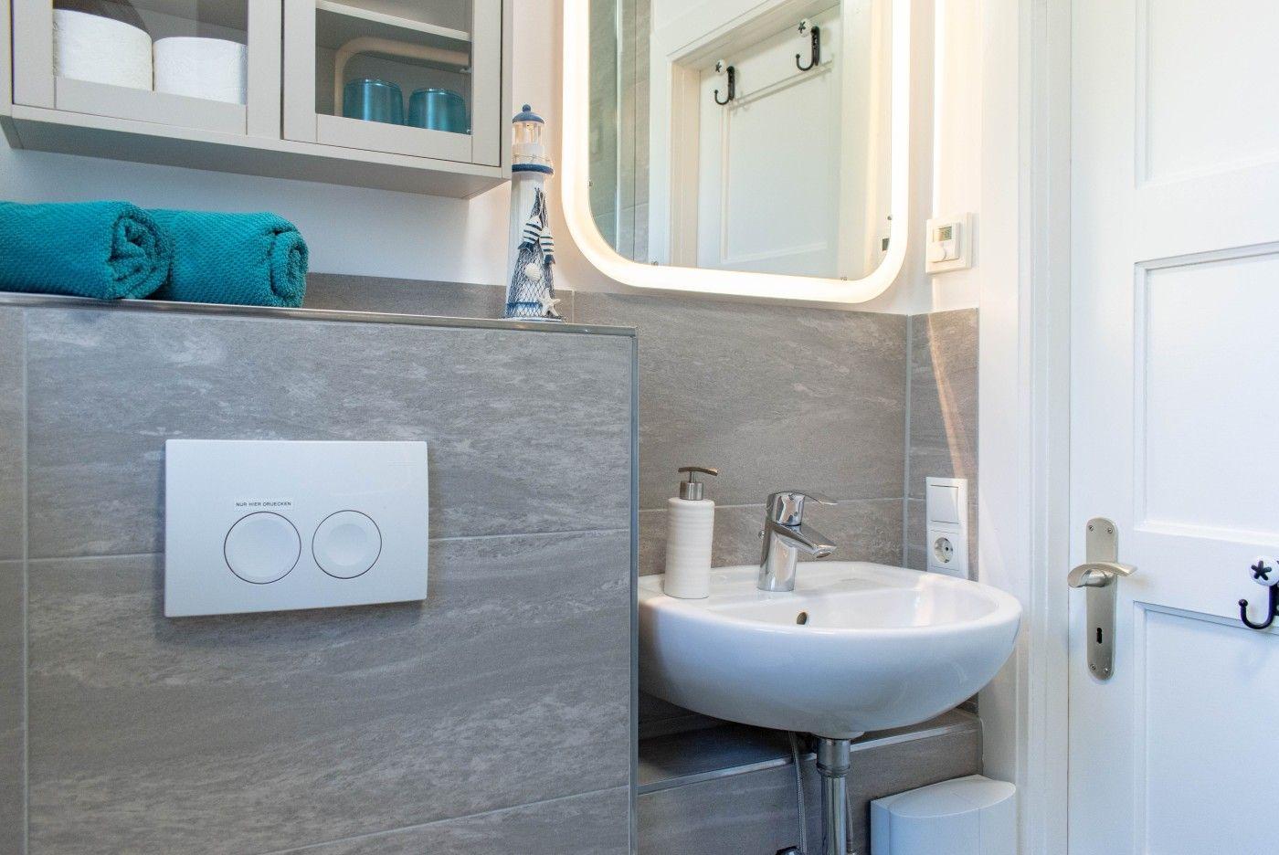 Badezimmer mit Waschbecken, Spiegel und Toilette. Aufgehängt sind Handtücher und ein Leuchtturm-Objekt.