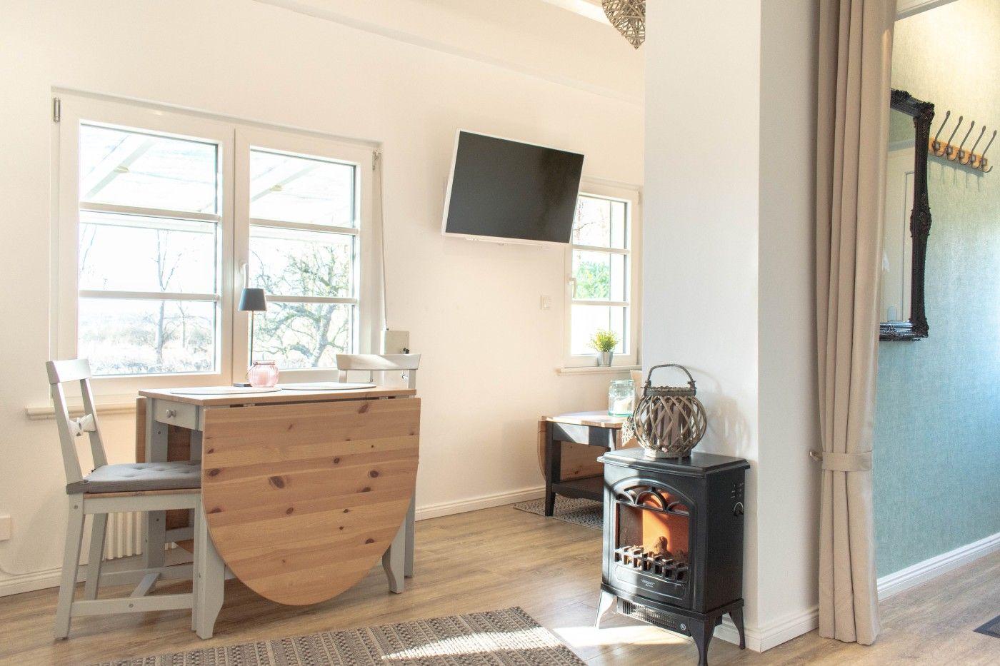 Ein Raum mit Esstisch, Fernseher und Kaminofen. Durch Fenster fällt Tageslicht.