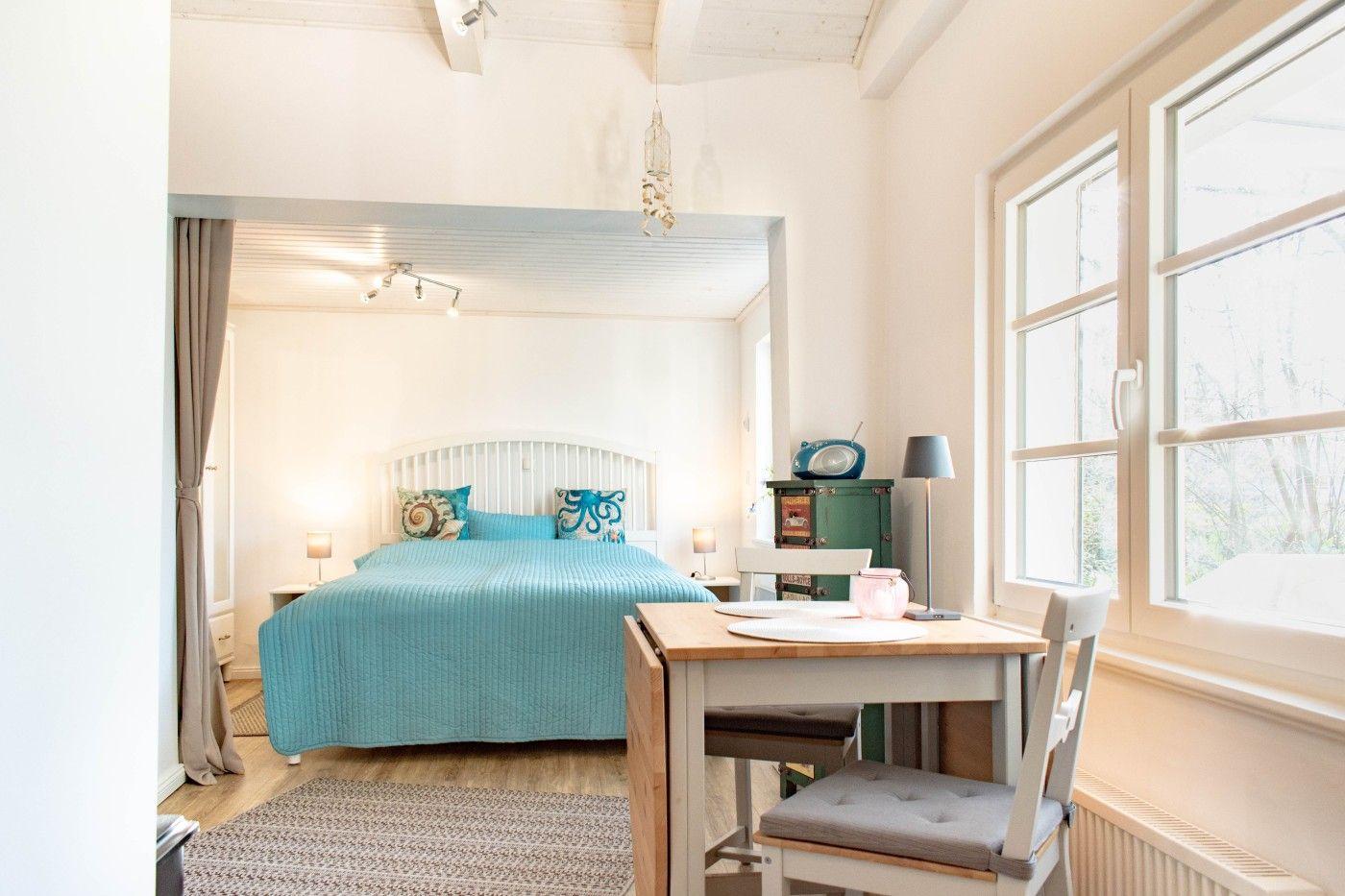 Schlafzimmer mit blauer Bettdecke, Schreibtisch und großen Fenstern.