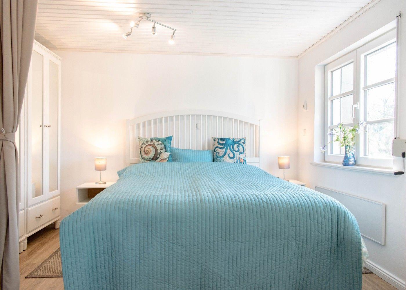 Schlafzimmer mit Bett, Nachttischen und Fenster. Blaue Bettwäsche und weiße Möbel.