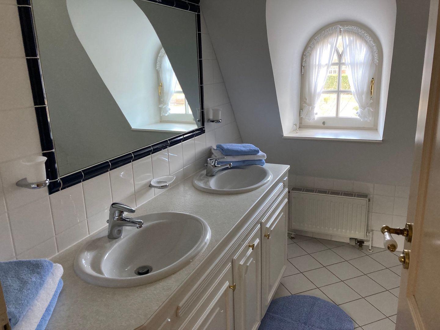 Badezimmer mit zwei Waschbecken, Spiegel und Fenster mit weißen Vorhängen.