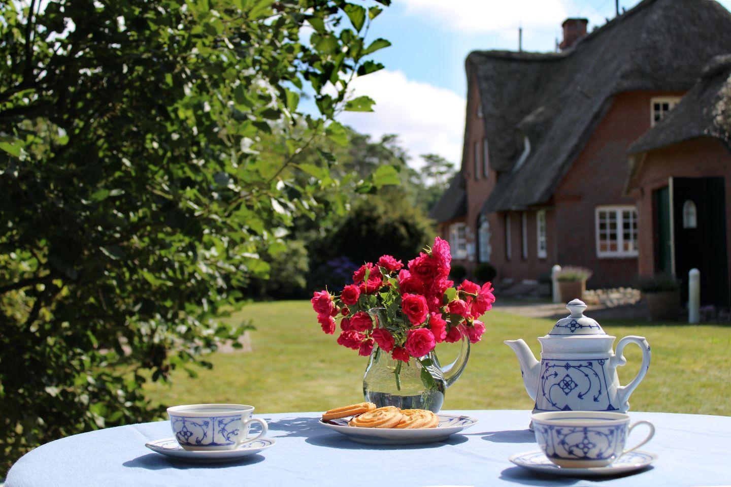 Tisch mit Teeservice, Rosen und Keksen vor einem Haus mit Strohdach.