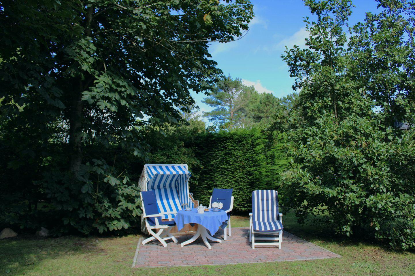 Außenbereich mit blau-weißen Strandkörben und Tisch auf Steinplatten umgeben von Bäumen.