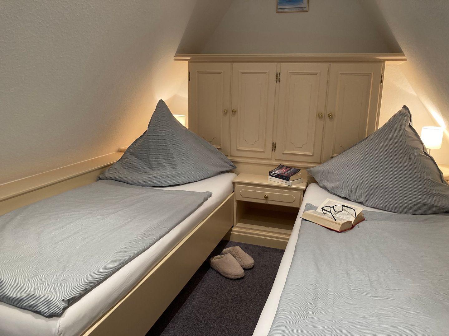 Zwei Betten mit grauen Bettwäschen, Nachttisch und Schrank unter Dachschrägen.