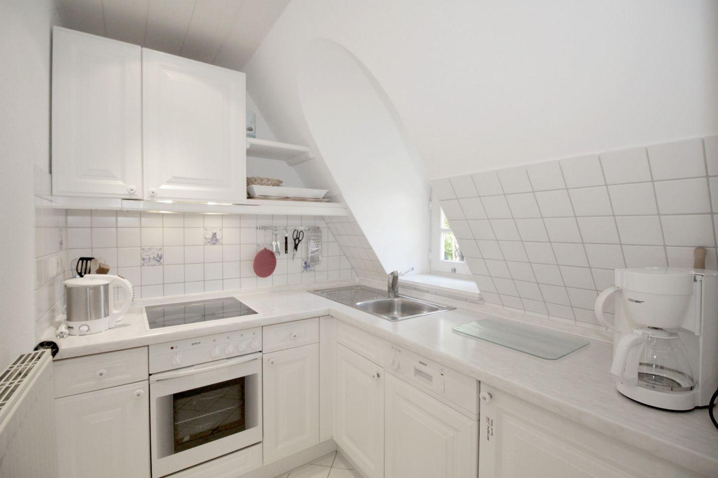 Küche mit weißen Schränken, Edelstahlspüle und Kochfeld. Dachschrägen Decke und Fenster.