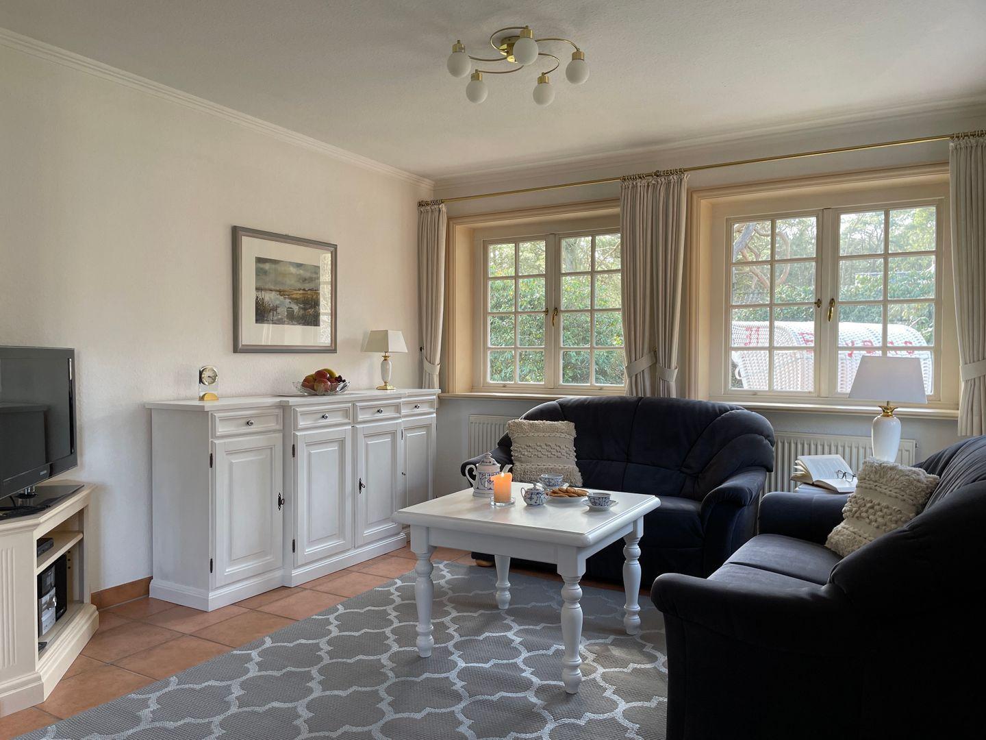 Wohnzimmer mit zwei dunklen Sofas, weißem Tisch und Fenstern mit Vorhängen.