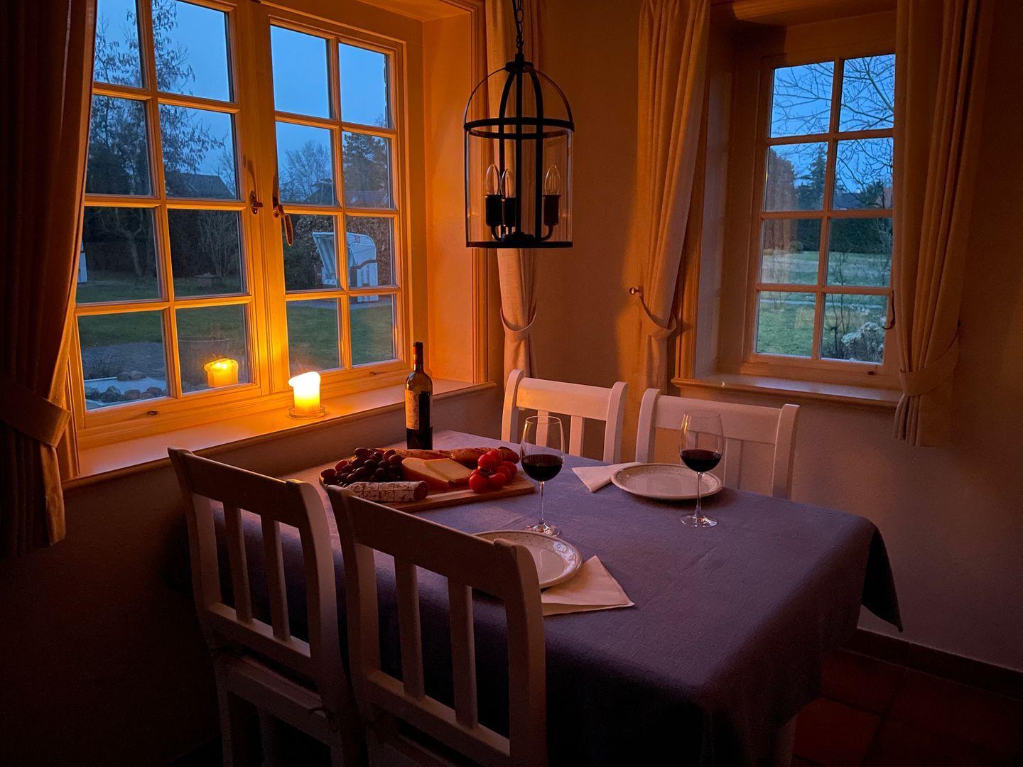 Esstisch mit Tischdecke, Stühlen, Wein und Obst vor Fenstern mit Blick auf Garten.