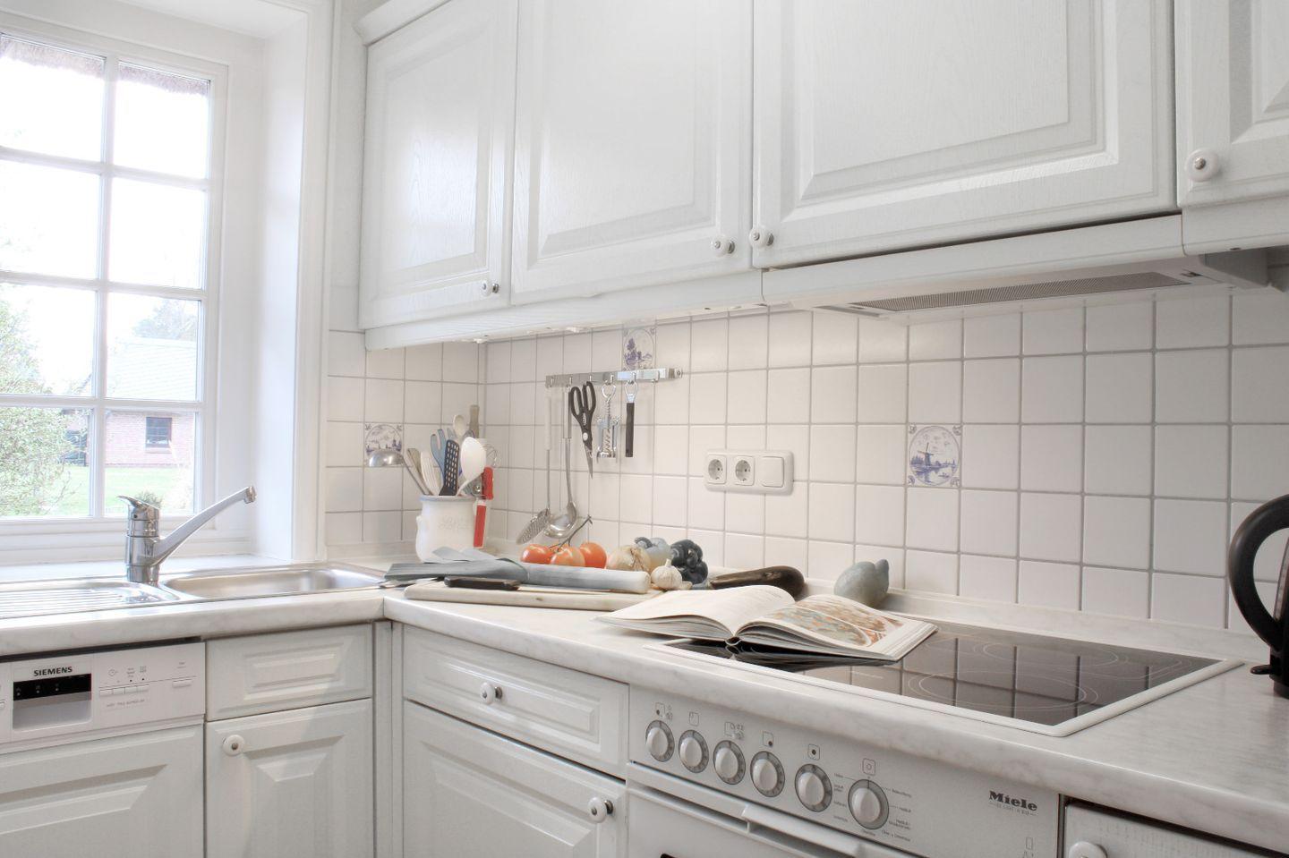 Küche mit weißen Schränken, Arbeitsfläche und Herd. Fenster, Spüle und Küchenutensilien sind sichtbar.