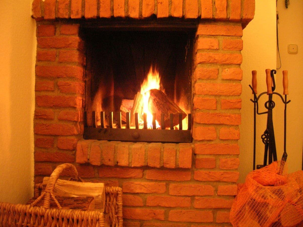 A fireplace with burning wood, surrounded by red bricks and a basket with firewood.