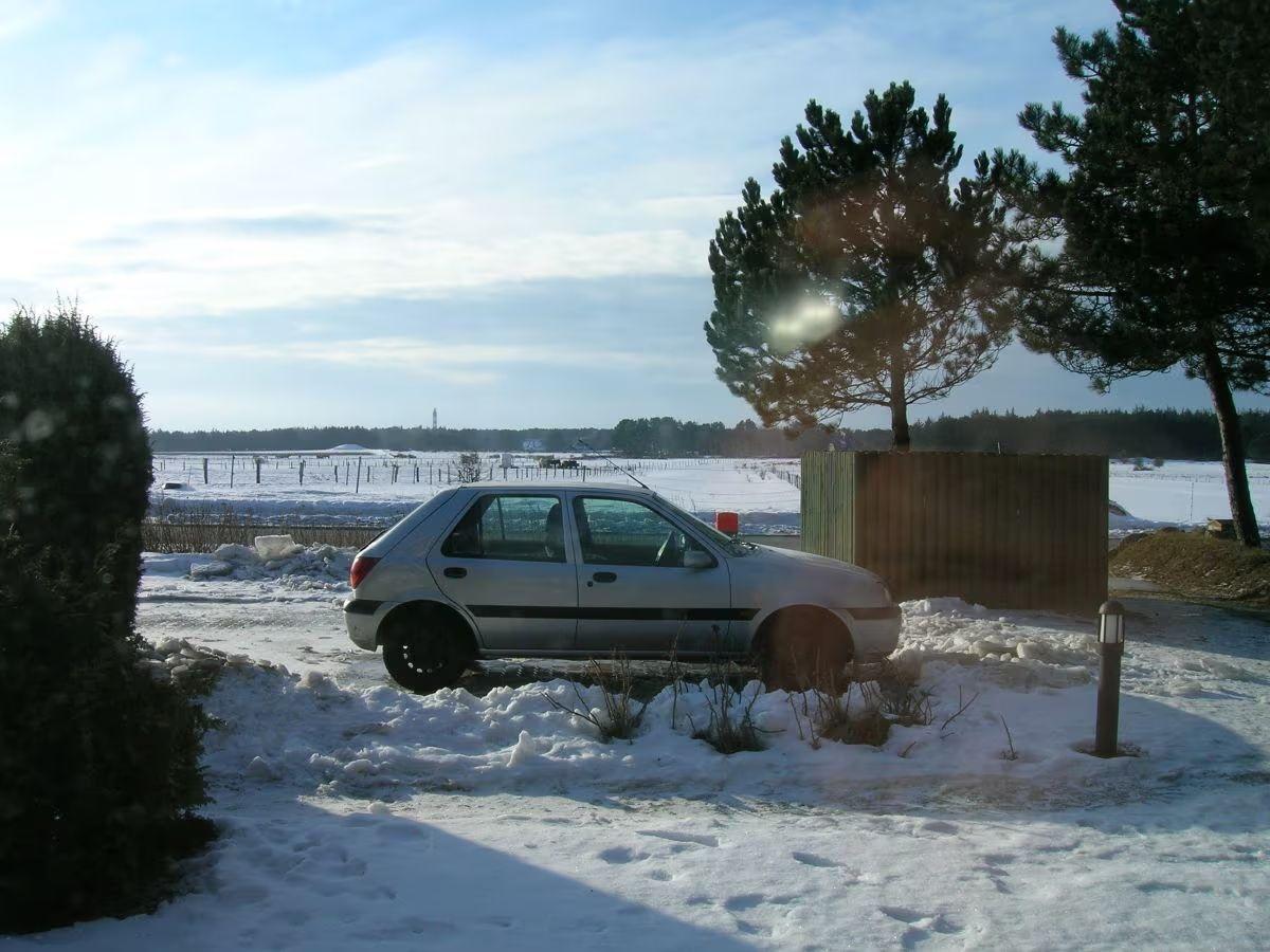 A silver car is parked on a snow-covered property beside trees and a fence.