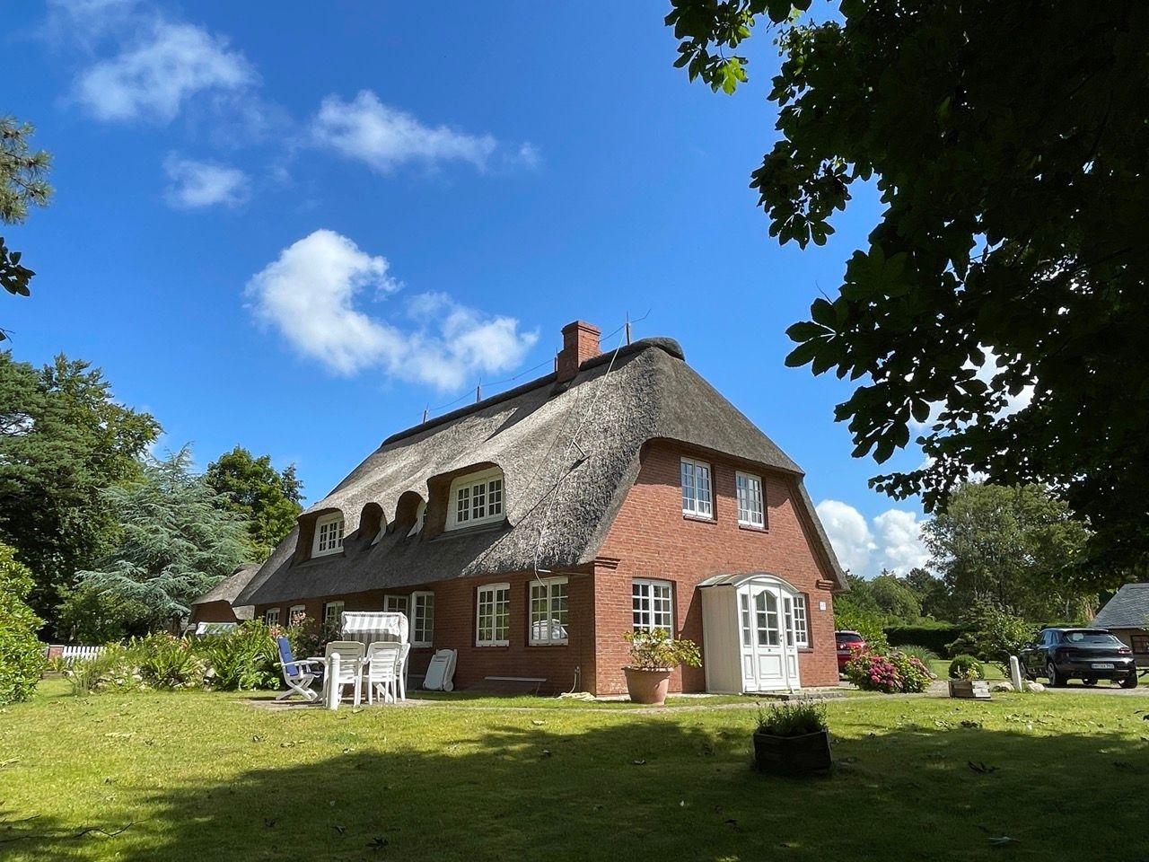 Rotes Backsteinhaus mit Strohdach und weißem Eingang. Grünflächen, Bäume und Gartenmöbel.